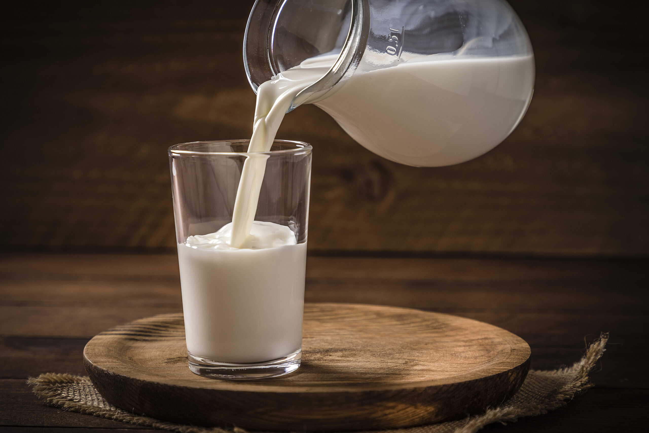 Front view of a pitcher pouring milk into a drinking glass on a rustic wooden background. Low key DSLR photo taken with Canon EOS 6D Mark II and Canon EF 24-105 mm f/4L