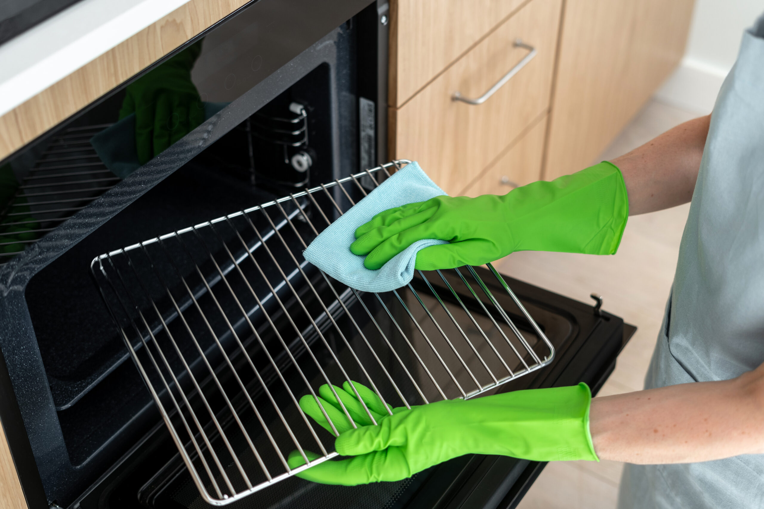 High angle view of woman in rubber gloves cleans the grill grid inside oven. Female using a cloth to wipe household appliance inregrated in kitchen. Housework and cleaning service concepts