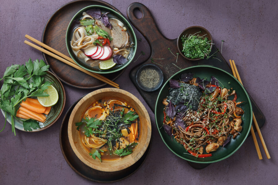 Japanese Ramen Noodle Soup and Udon Noodle Soup with Miso. Yaki Soba noodles with chicken and vegetables. Flat lay top-down composition on dark purple background. Horizontal image.