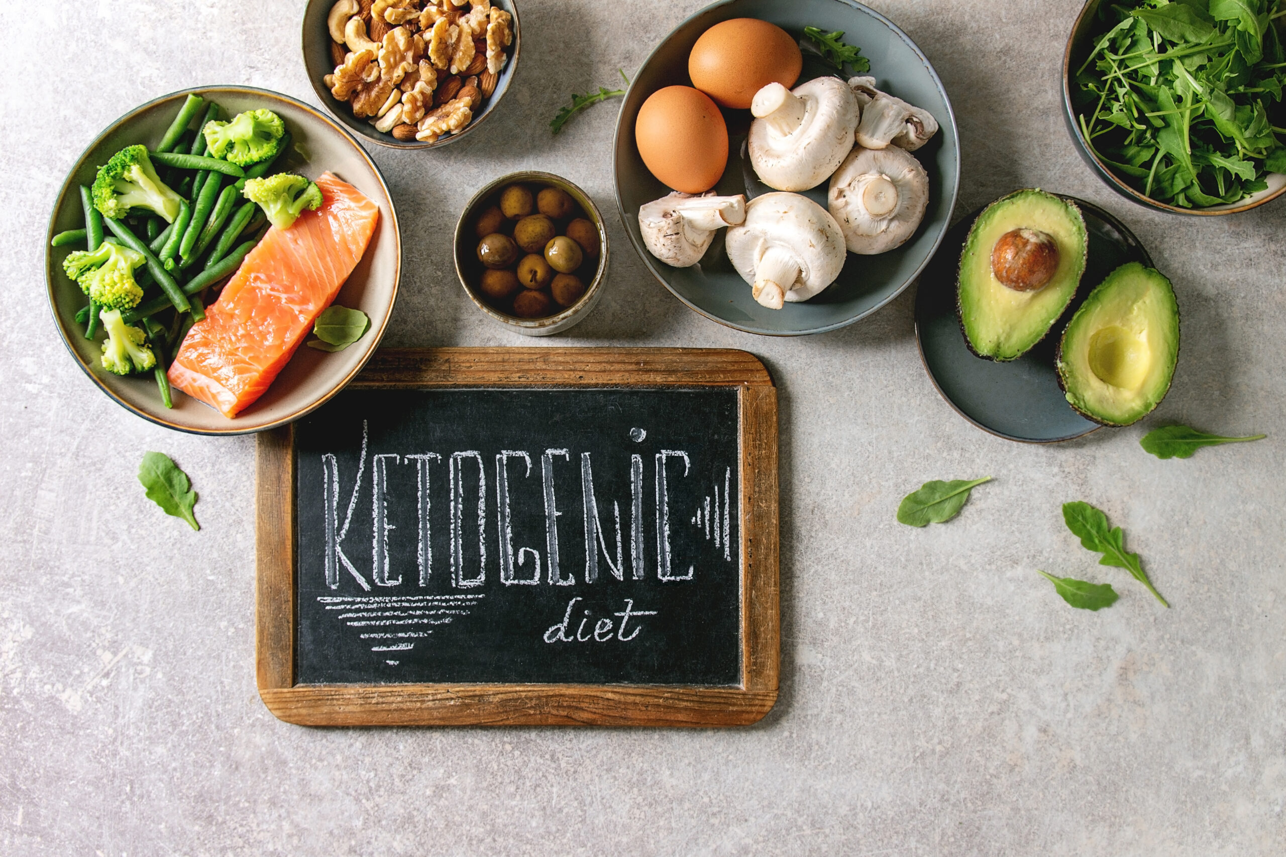 Ketogenic diet ingredients for cooking dinner. Raw salmon, avocado, broccoli, bean, olives, nuts, mushrooms, eggs in ceramic bowls with chalk board lettering. Grey texture background. Flat lay, space