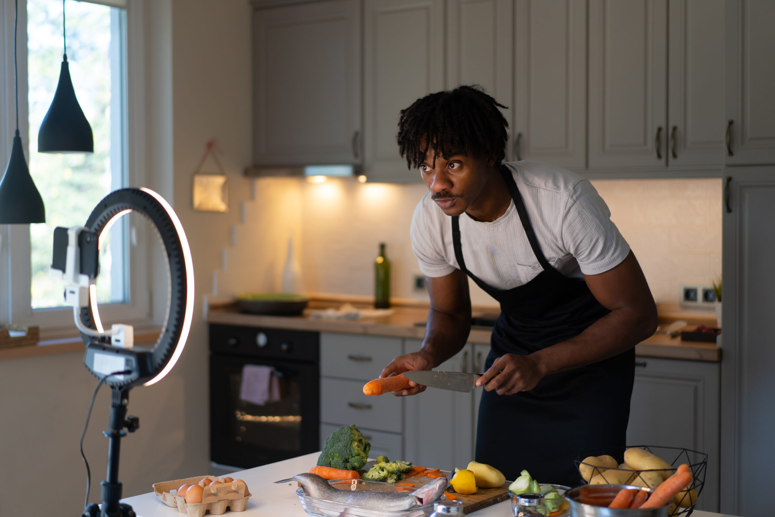 LED ring lamp supporting a mobile phone to record a live streaming event on cooking, an African-American man showing an easy healthy meal from his kitchen at home