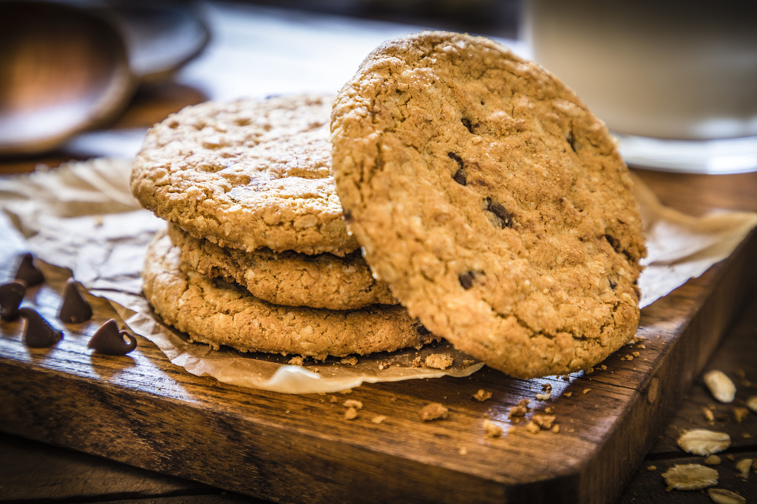 Pile of homemade oatmeal cookies with chocolate chips upon a cutting board on rustic wooden table. Cookies are surrounded by oat flakes, oat spikes and chocolate chips. Main focus is on cookies and in the defocused background is a glass full of milk. Low key DSLR photo taken with Canon EOS 6D Mark II and Canon EF 24-105 mm f/4L