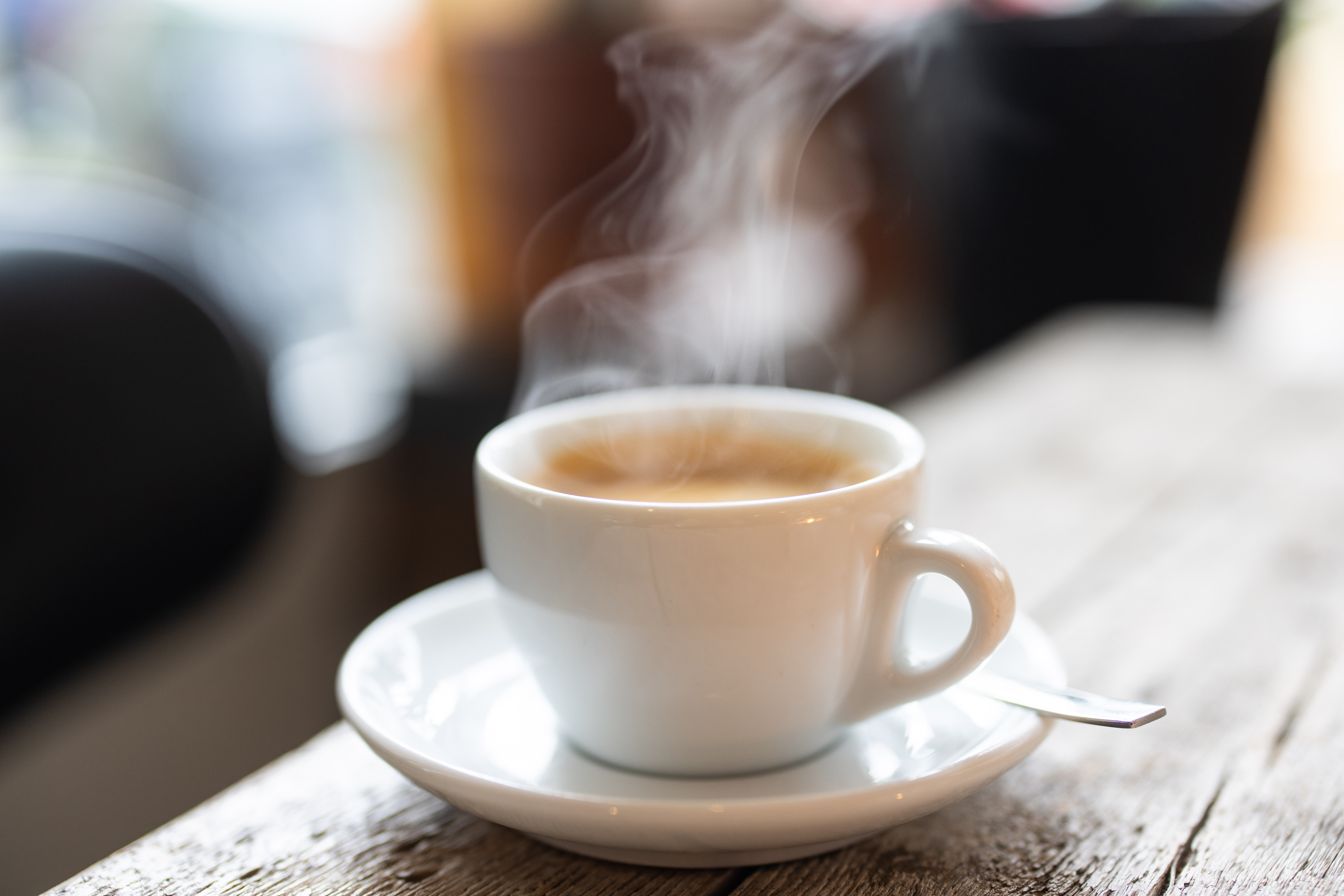 Steam rising from a white cup of hot coffee with a spoon on a saucer over a wooden table in the cafe. Close-up of a refreshing hot cup of a coffee at a cafe.