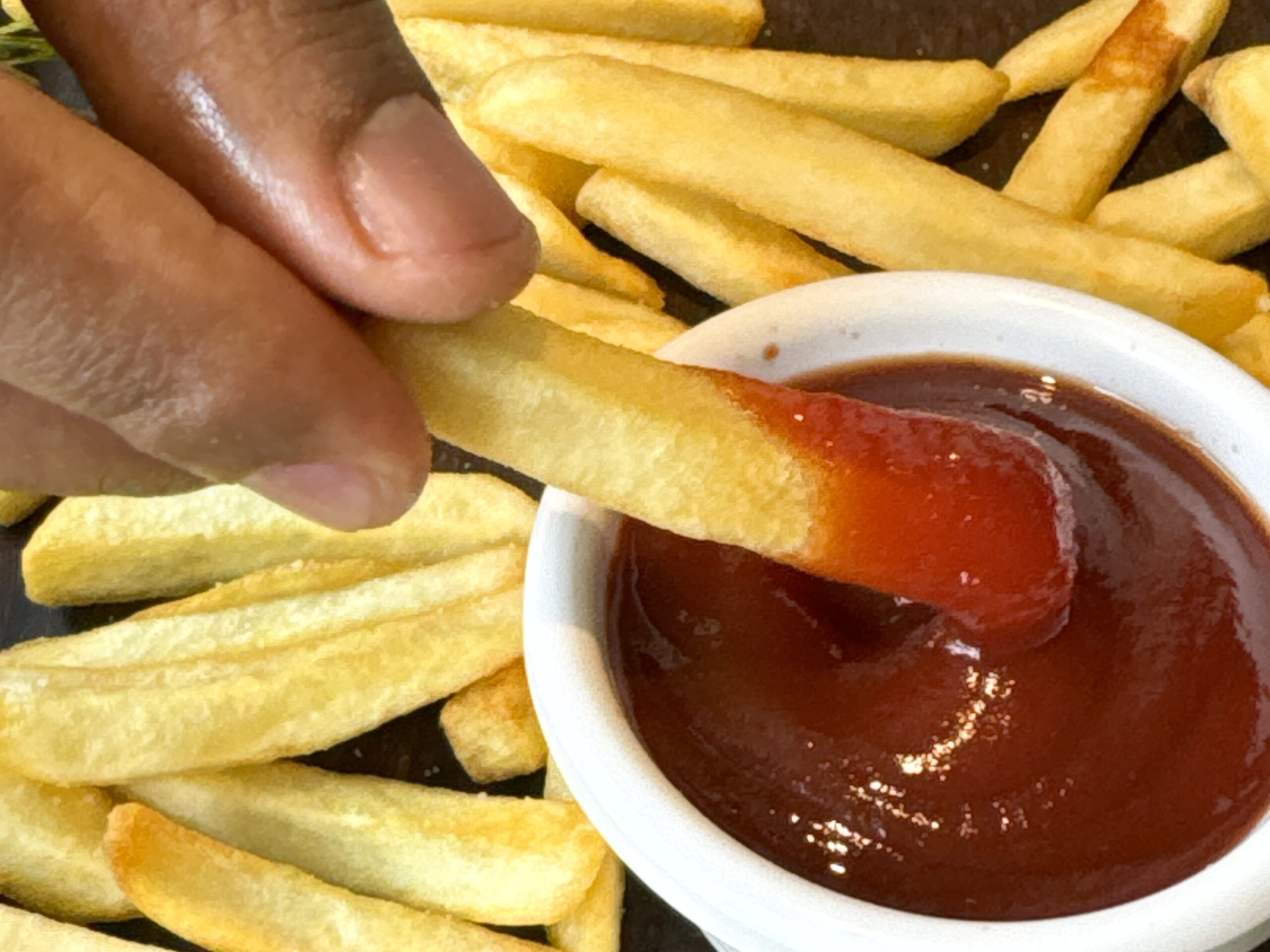 Stock photo showing close-up, elevated view of pile of crispy, golden, French fry chips and bowl ketchup of on wooden table.