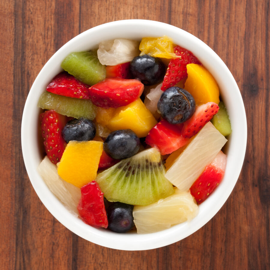 Top view of white bowl full of fruit salad containing strawberries, blueberries, orange, kiwi, pineapple and peach