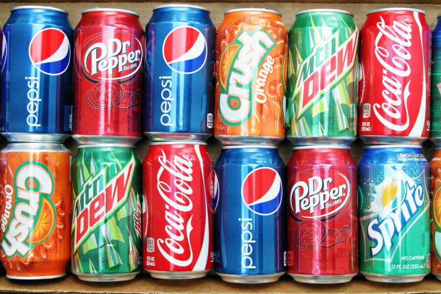 Variety of brand name soda cans placed in a box in a double row. Brands include Coca Cola, Pepsi, Dr Pepper, Orange Crush, Sprite and Mountain Dew.