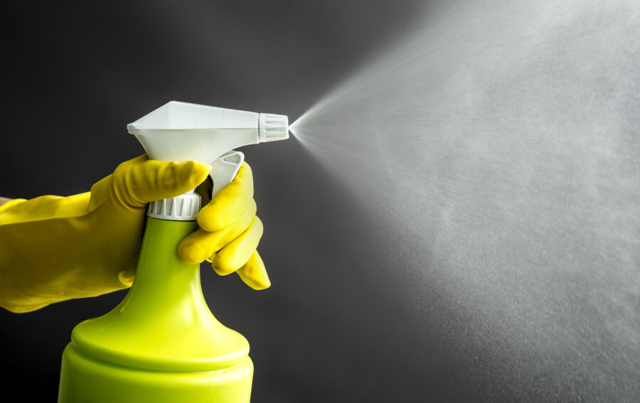 Woman wearing yellow rubber gloves using green spray bottle and spraying liquid mist in air, cool lighting effect. Lot of copy space.