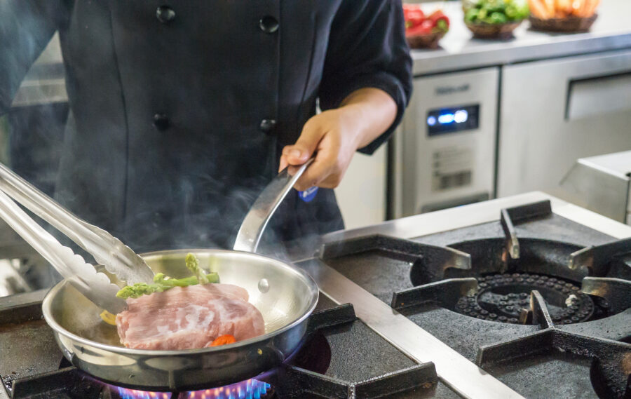 Close-up view of a woman's torso wearing a black uniform and her hand as she cooks food in a frying pan on a gas burner of an industrial cooking stove top in a professional kitchen where stainless steel appliances are visible in the background.