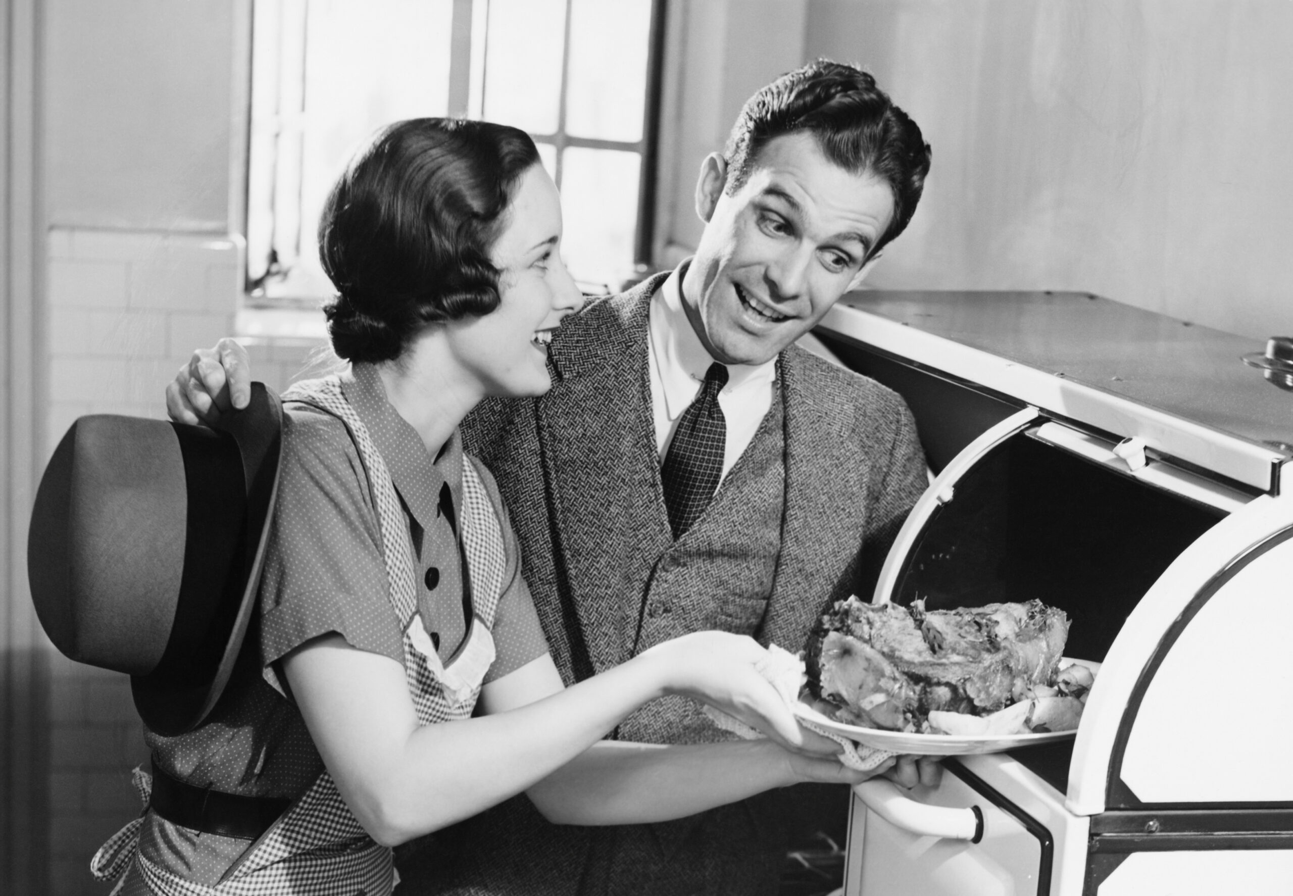 Couple in kitchen, wife taking roast from oven, (B&W)