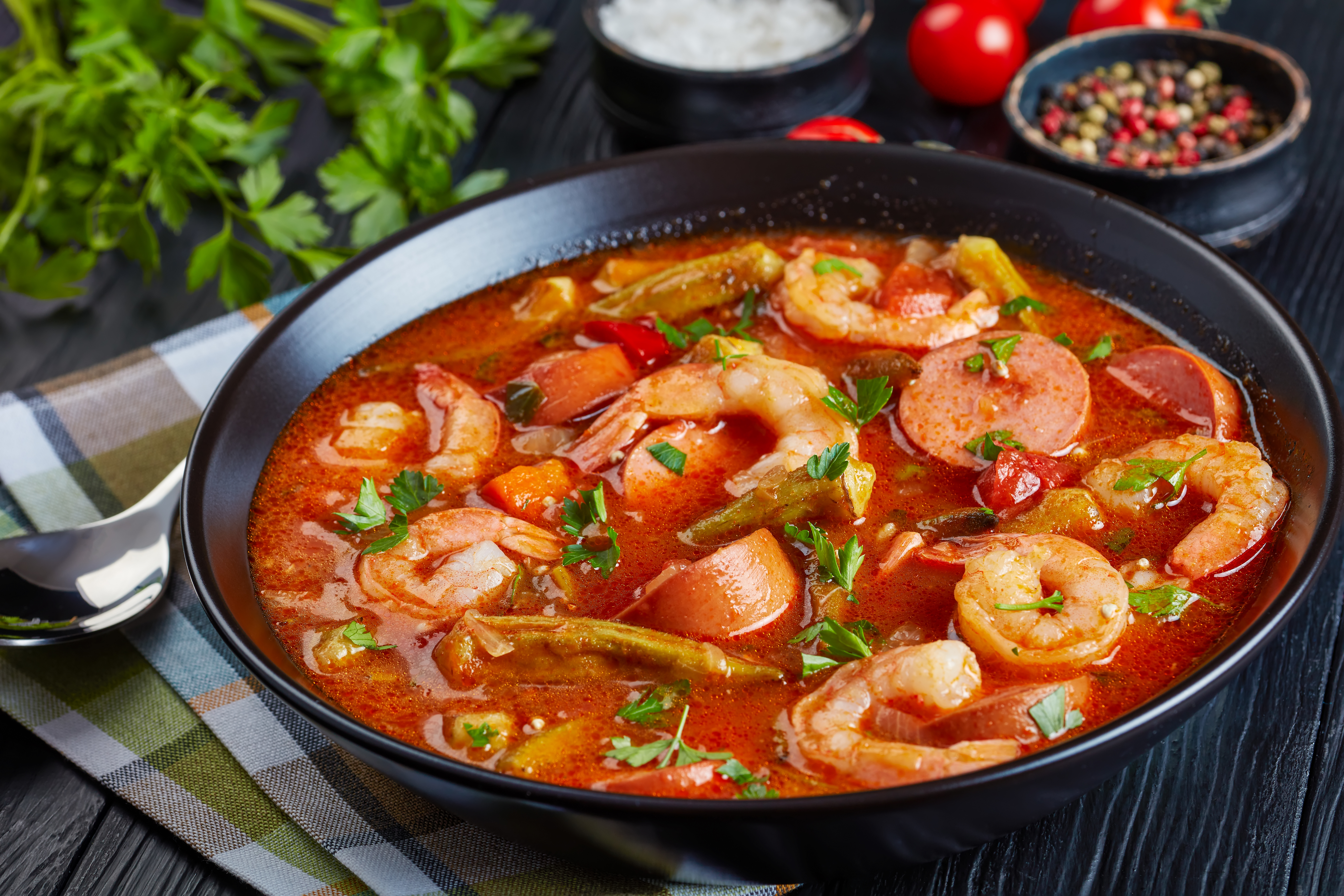 gumbo with prawns, baby okra and sausage in a bowl on a black table with napkin and silver spoon, view from above, close-up