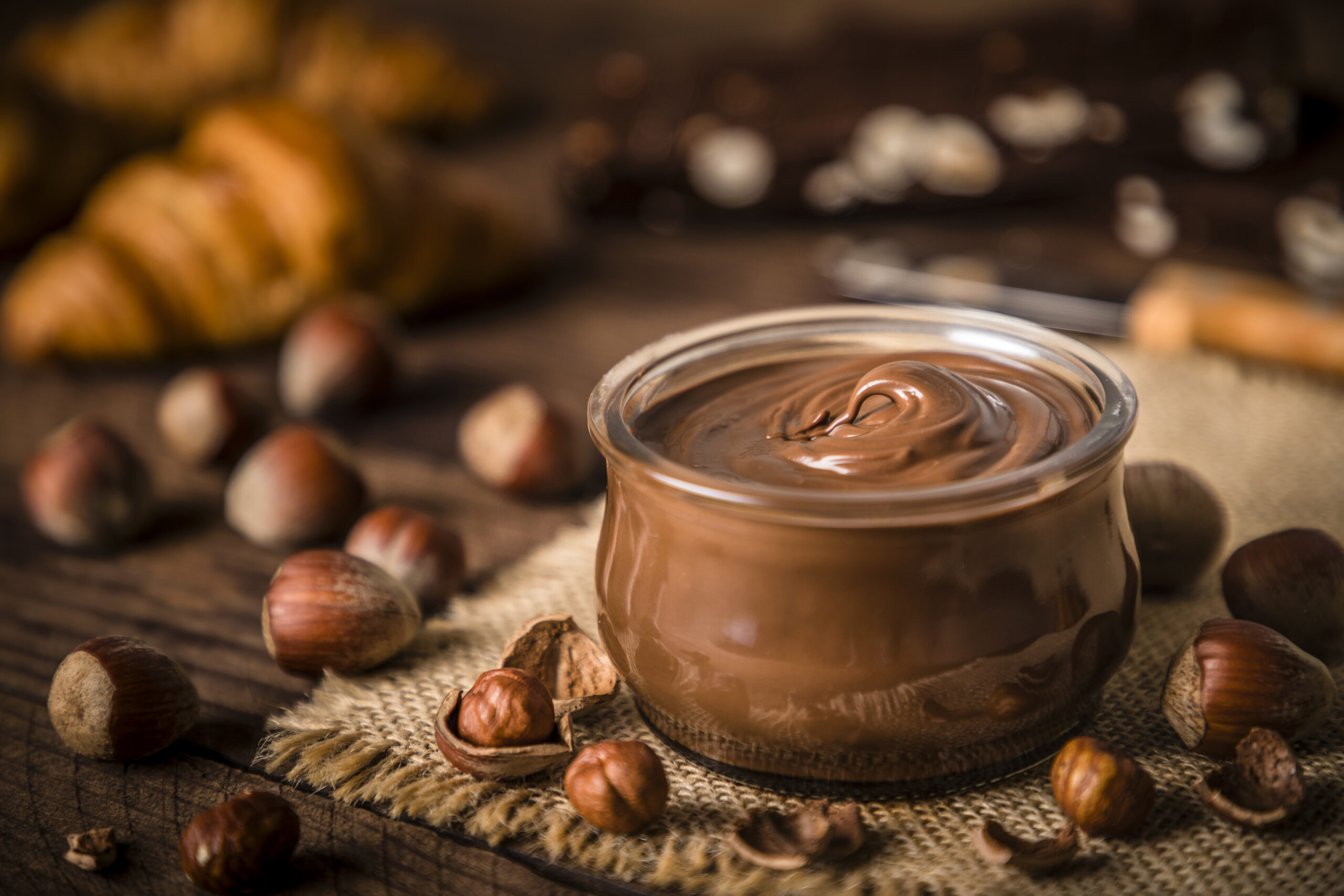 Front view of a crystal jar full of chocolate and hazelnut spread surrounded by some hazelnuts on a rustic wooden table. Selective focus is on the jar and on the defocused background are some croissants and chocolate bars. Predominant color is brown. Low key DSLR photo taken with Canon EOS 6D Mark II and Canon EF 24-105 mm f/4L