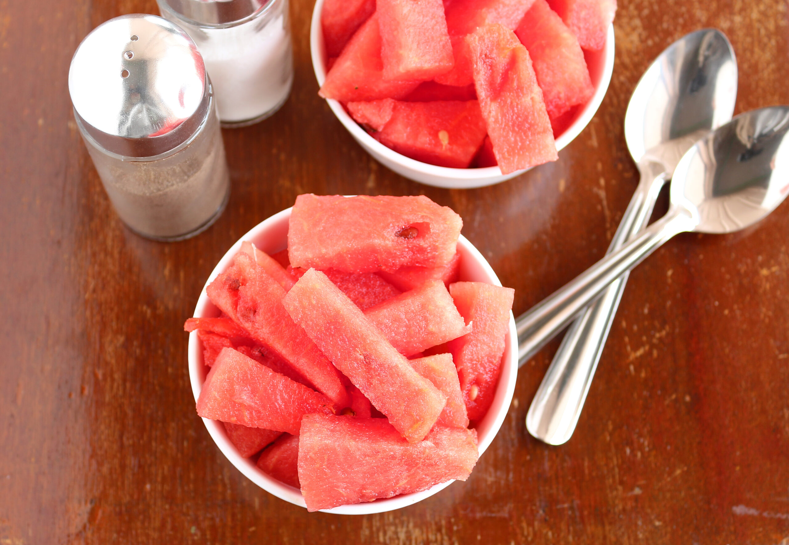 Healthy snack watermelon in bowls and bottles of salt and pepper and steel spoons on a wooden table.