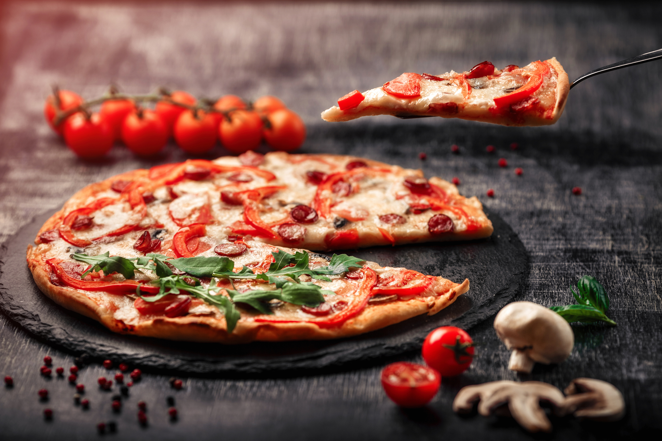 A slice of pizza on a spatula with smoked sausages, cheese, mushrooms, cherry tomatoes, bell peppers and greens on a stone and a black chalk board.