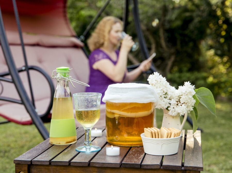 Selective focus on jar with kombucha drink and tea mushroom in it and served in goblet glass outdoors in sunny summer evening, blurred tea drinking woman on the background.