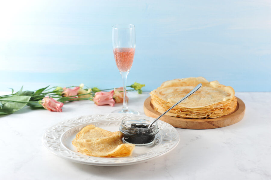 Thin pancakes on a wooden tray. Next to the plate with pancakes and black caviar. In a cup of caviar a teaspoon. In the background, pink flowers Eustoma and a glass of pink champagne.