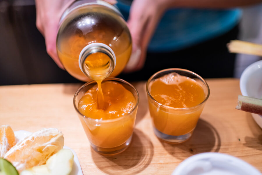 person serving pouring kombucha fermented tea into two juice shot glasses on wooden table from bottle
