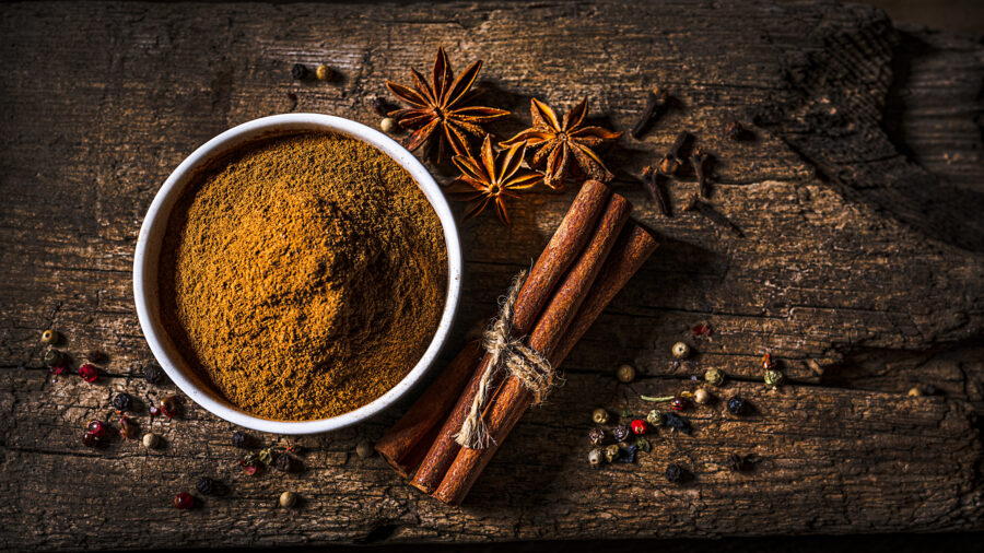 top view of a white bowl filled with cinnamon powder shot on rustic wooden table. Cinnamon sticks, star anise, cloves and peppercorns are all around the bowl.  Predominant color is brown. The composition is at the left of a horizontal frame 