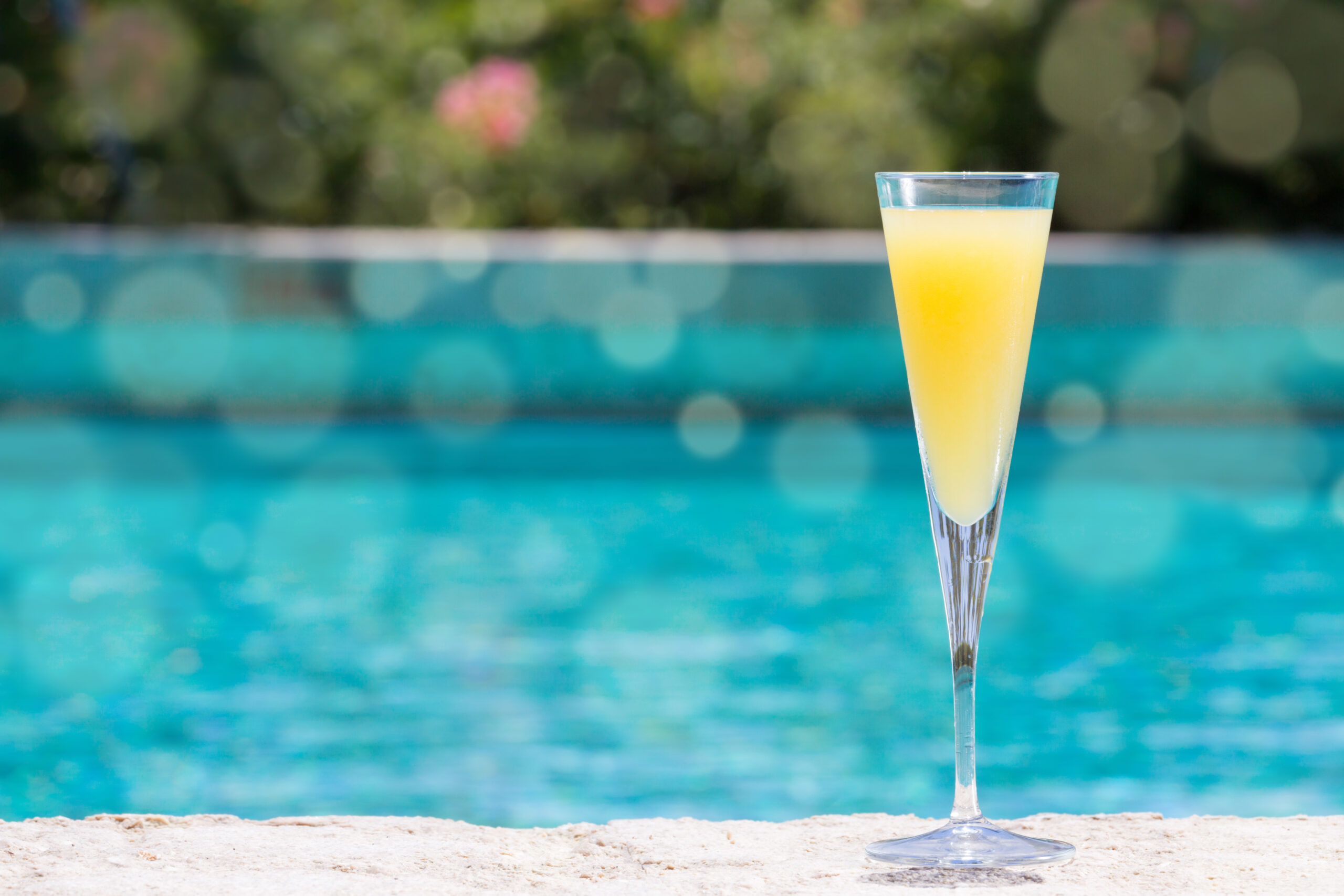 Glass of Mimosa cocktail on the pool nosing at the tropical resort. Horizontal, cocktail on right side. Bokeh details