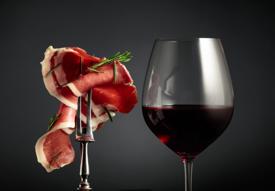 Glass of red wine and sliced prosciutto with rosemary on a fork, black background.