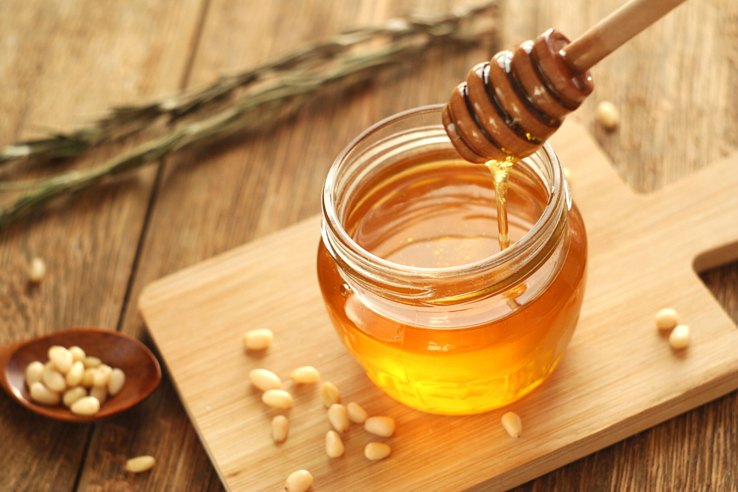 Honey is in a jar on a wooden table. Honey flows down the honey stick into the jar. Nearby are pine nuts and rosemary. selective focus