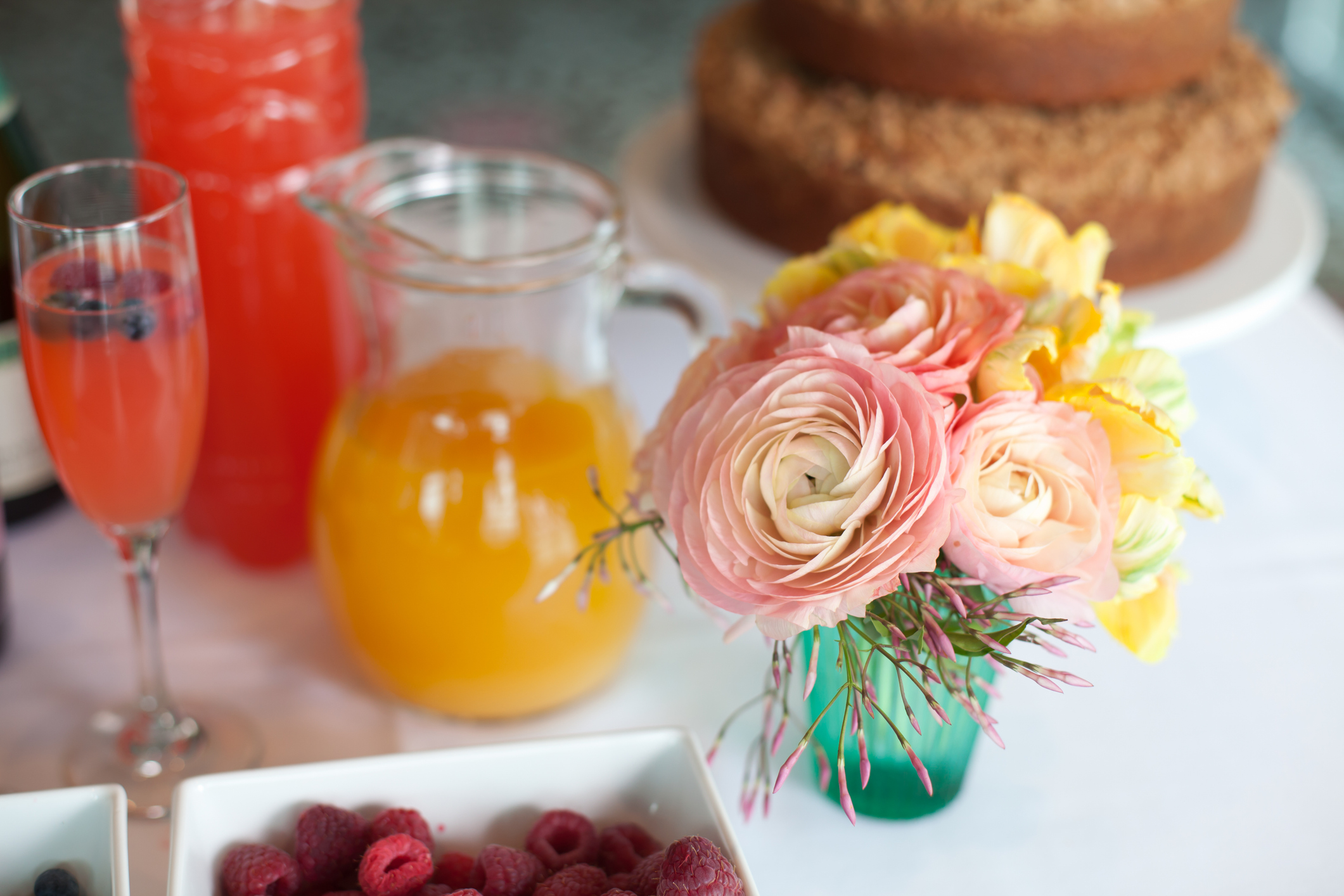 "An arrangement of pink ranunculus flowers on a white breakfast table with coffee cake, mimosa, juice and berries."