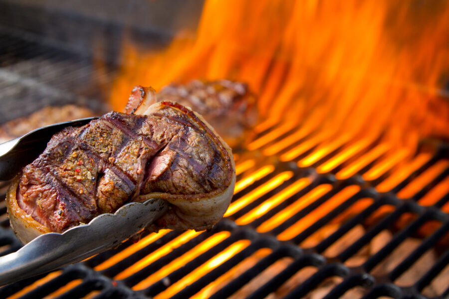 Thick bacon wrapped filet mignon cooking, grill marks held in tongs over a big fire in an old fashioned charcoal barbecue grill .