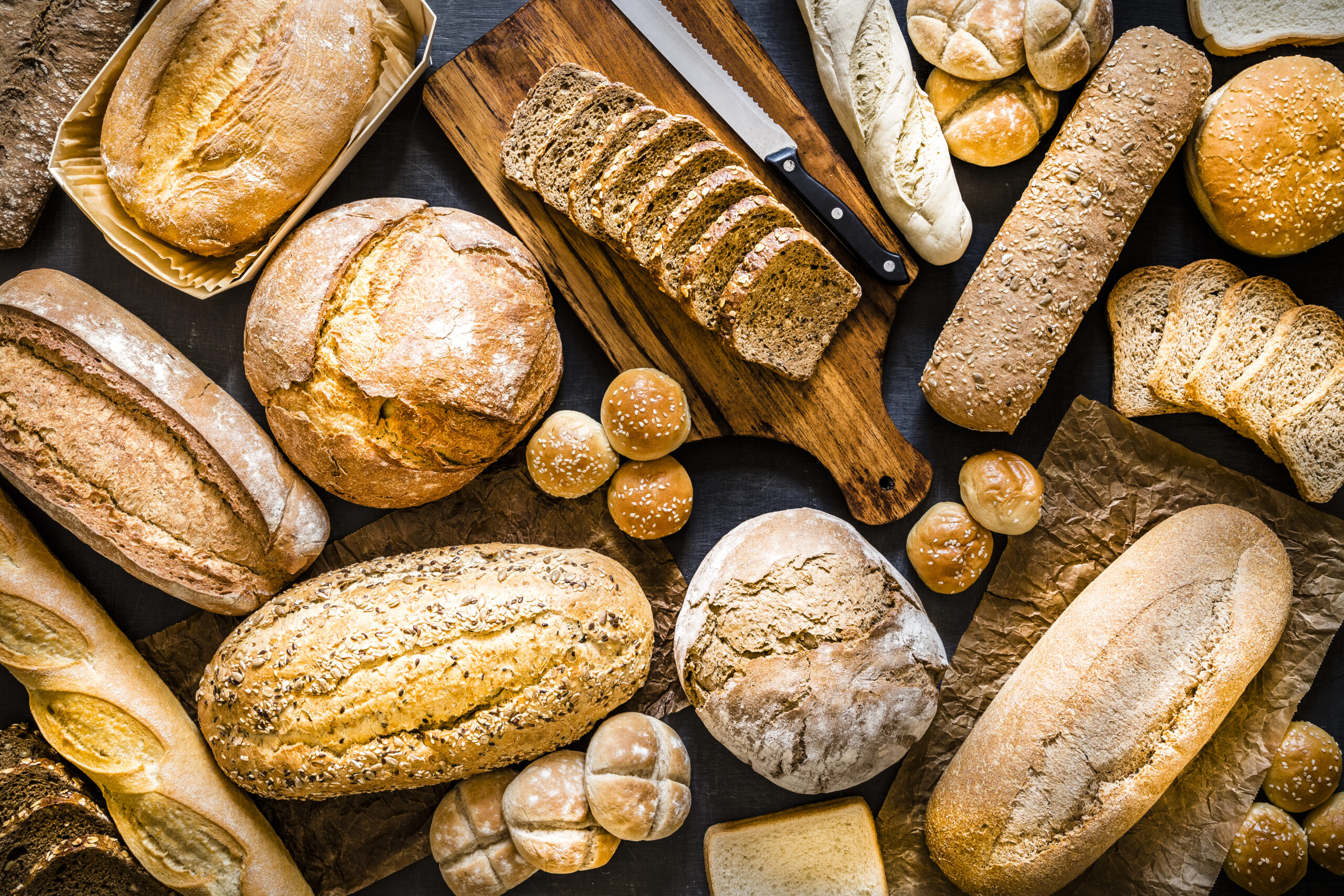 Top view of various kinds of breads like brunch bread, rolls, wheat bread, rye bread, sliced bread, wholemeal toast, spelt bread and kamut bread. Breads are scattered making a background. Low key DSLR photo taken with Canon EOS 6D Mark II and Canon EF 24-105 mm f/4L