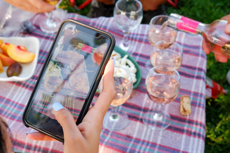 Woman's hand taking a photo of glasses of rose wine and cheese board with her smartphone on a picnic table.