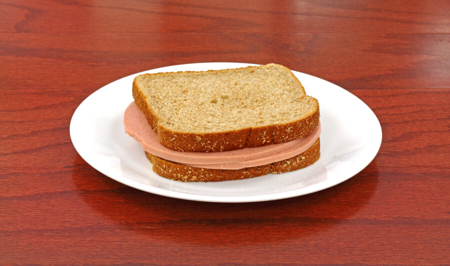 A bologna sandwich on wheat bread in a white dish on wood tabletop.
