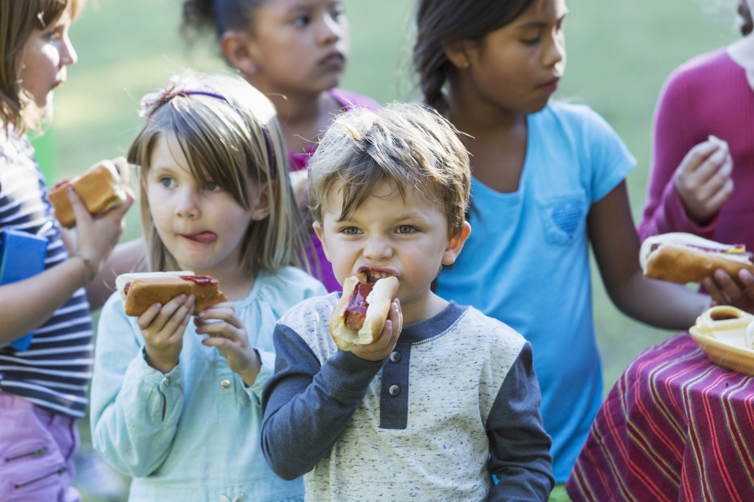 A little boy with a group of children eating hotdogs outdoors.  He is standing next to a little girl, taking a bite out of his hotdog.