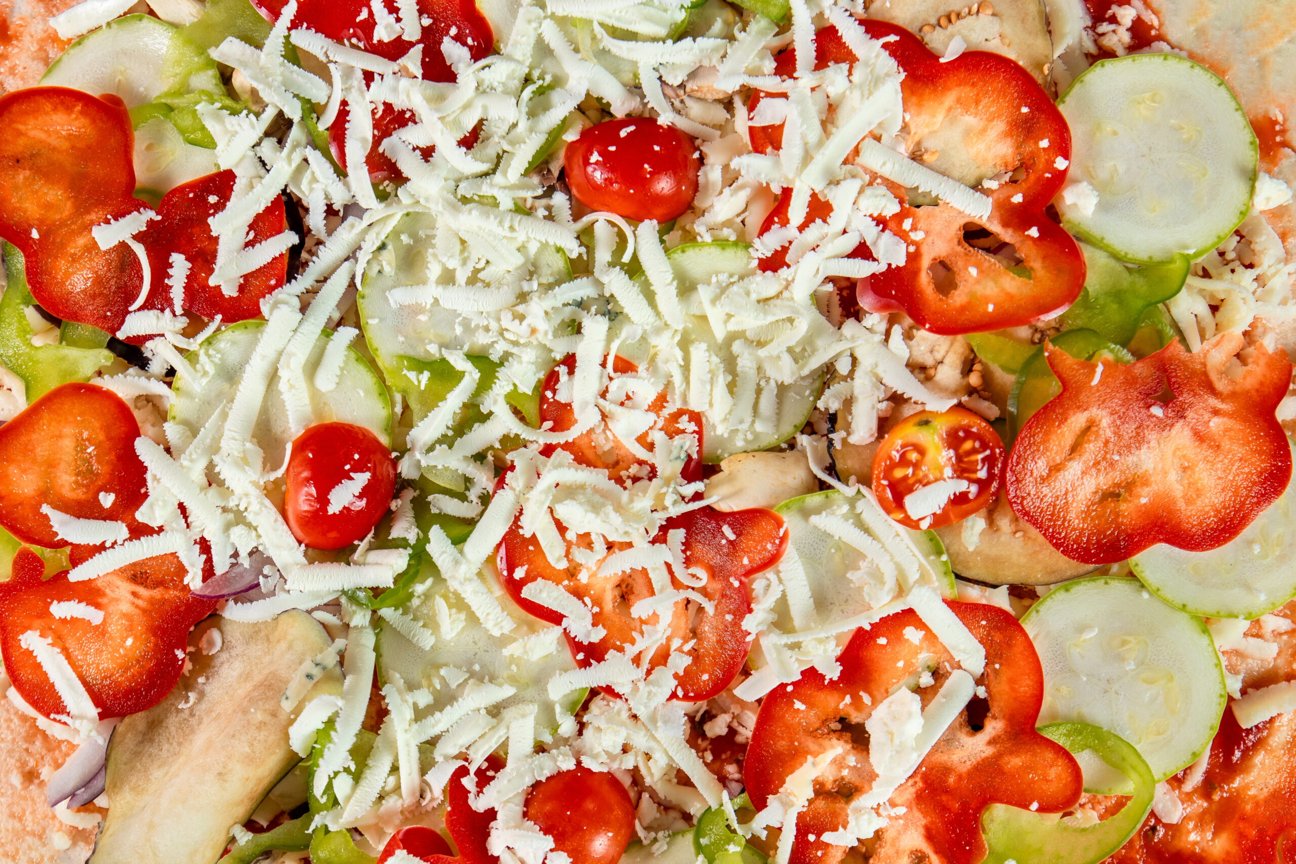 An up-close shot of a vegetarian pizza before baking, featuring mozzarella cheese, tomatoes, red and green bell peppers, and slices of cucumber, arranged beautifullyon the dough