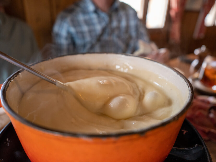 Bubbling cheese fondue in an orange cast iron pot. A long handled metal fondue fork with a lump of bread is submerged in the melted cheese, about to be lifted out and eaten. In the background a man in a checked shirt is sitting waiting for his turn.