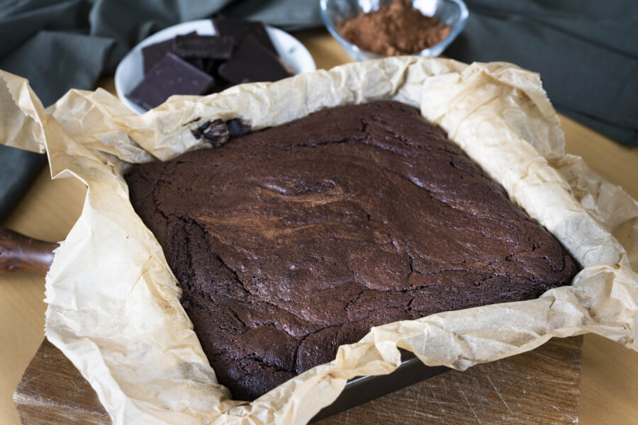 Chocolate brownie cake baked in a baking tin with parchment paper in the baking tin and cocoa powder and chocolate in the background.