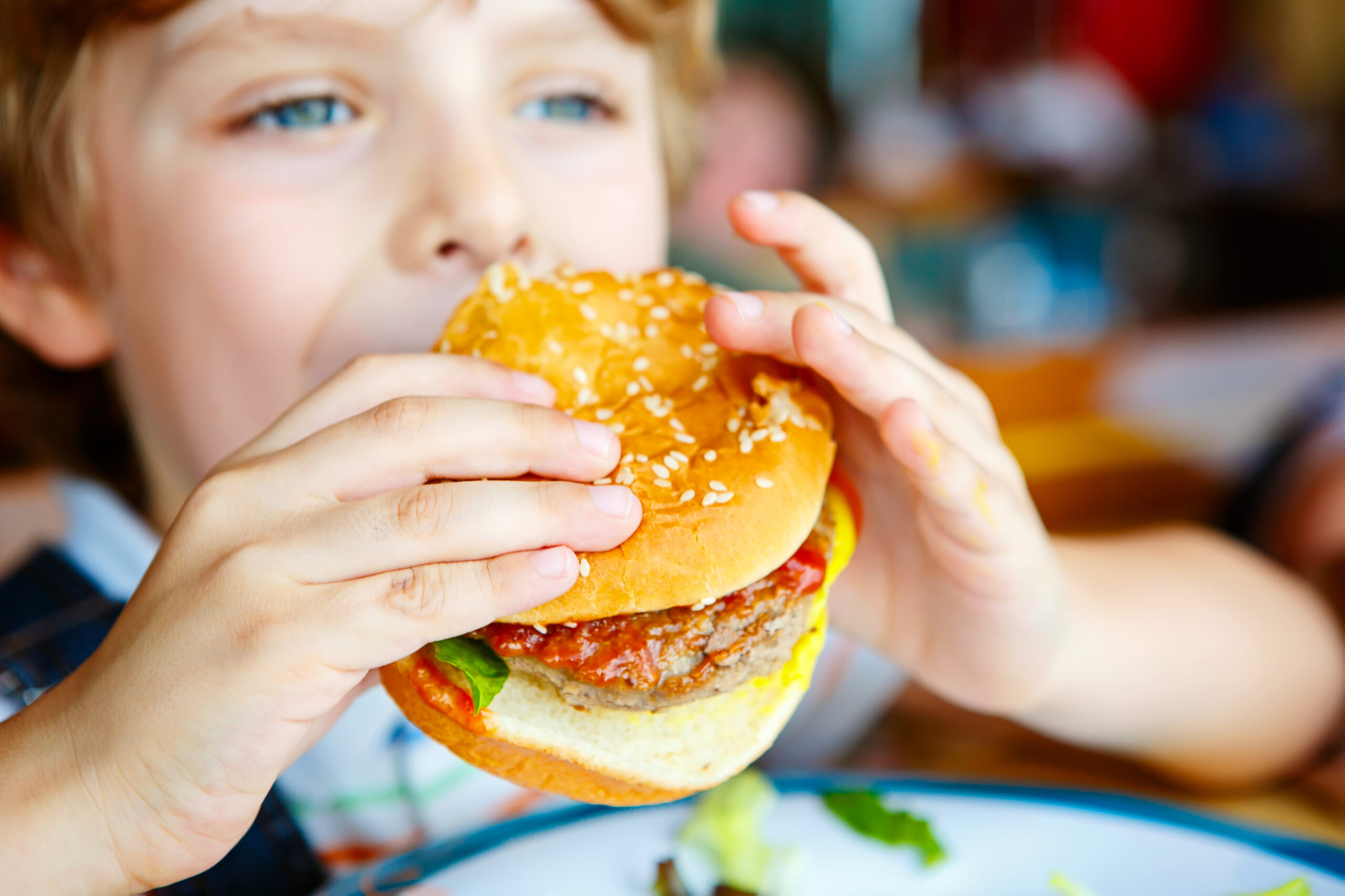 Cute healthy preschool kid boy eats hamburger sitting in cafe outdoors. Happy child eating unhealthy food in restaurant.