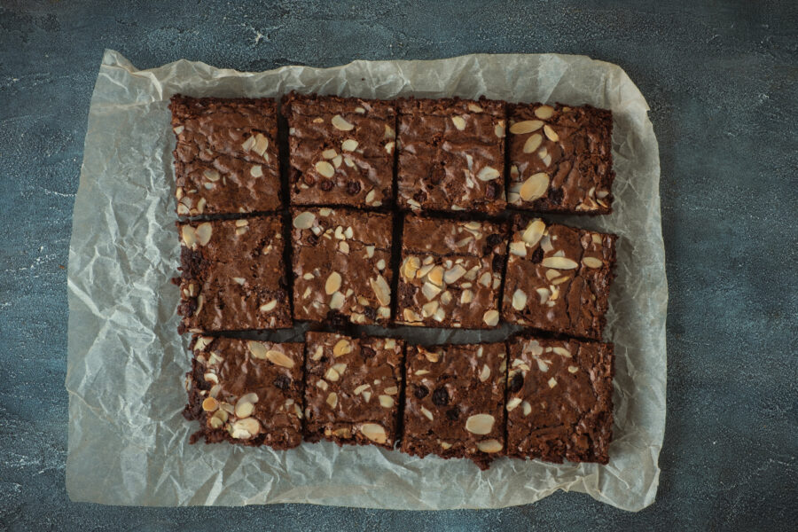 Delicious homemade chocolate brownies with almond flakes and raisins cut on square slices on the baking parchment. Flat lay. Grey concrete background