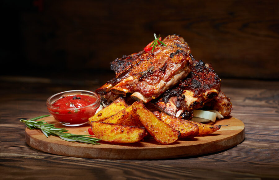 Fried ribs with rosemary, potatoes rustic, onion, sauce on wooden round Board. Dark background. 