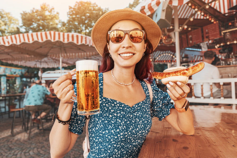 Happy girl drinking beer and eating traditional german bratwurst - hotdog at funfair and street food festival. 