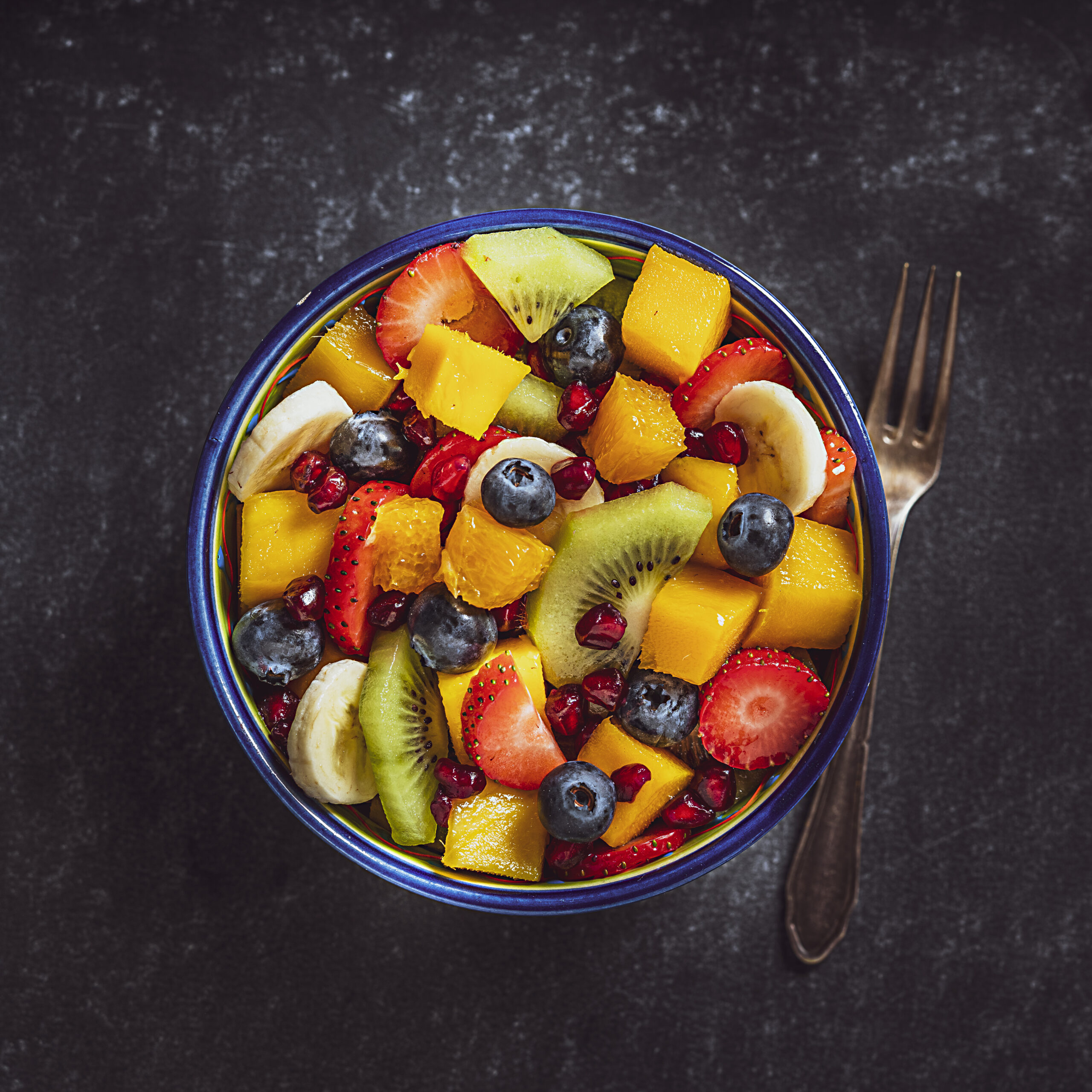 Healthy eating: fresh colorful homemade salad bowl shot from above on dark table. A fork is beside the bowl. Fruits included in the salad are mango, orange, kiwi, strawberry, banana, pomegranate and blueberries. High resolution studio digital capture taken with Sony A7rII and Sony FE 90mm f2.8 macro G OSS lens