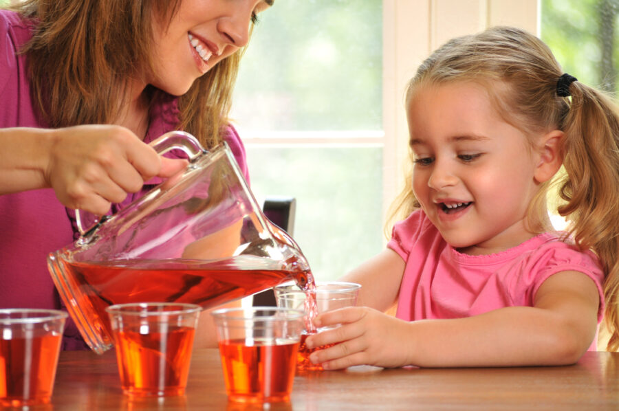 horizontal image of a young girl helping her mother with pouring drinks or juice, for a party or guests. the little girl has an enthusiastic expression. Focus in on the girl and a small amount of noise has been added to the background in post-processing.