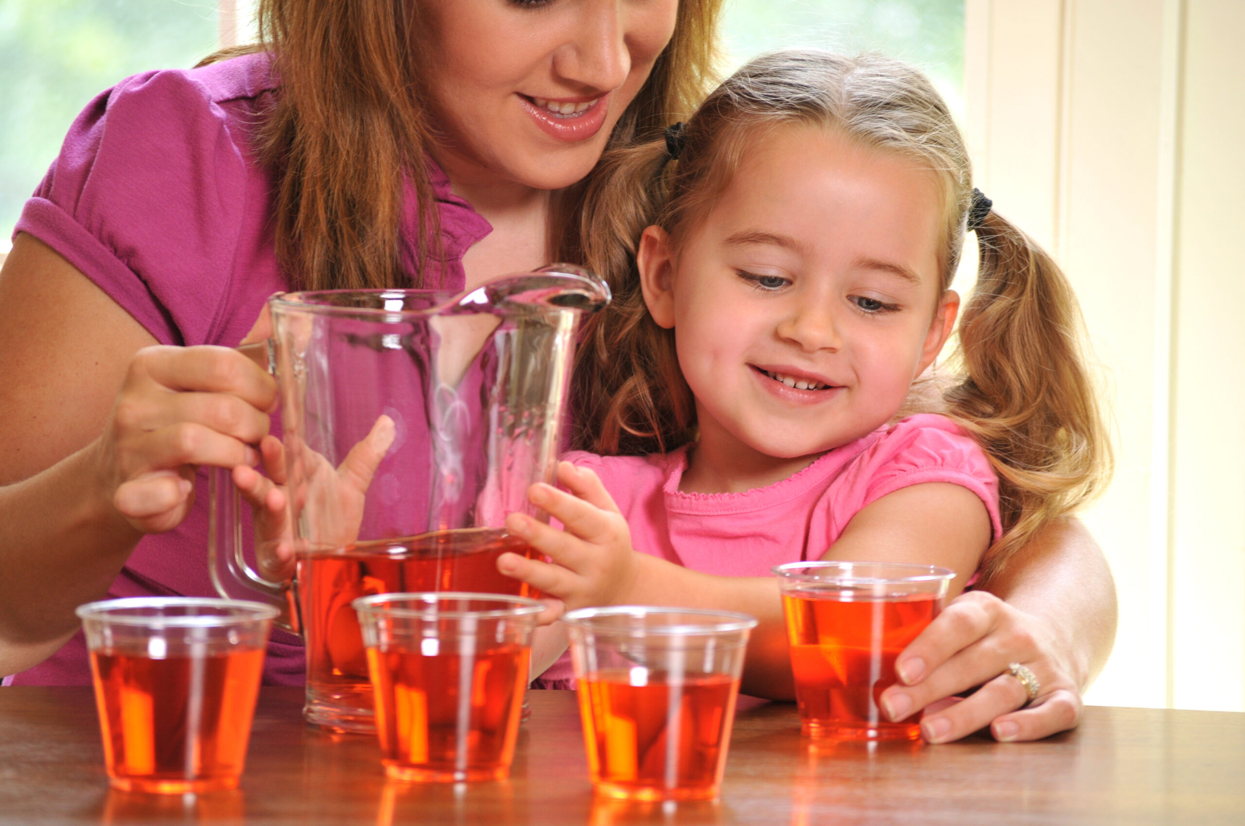 horizontal image of a young girl helping her mother with pouring drinks or juice, for a party or guests. the little girl has an enthusiastic expression. Focus in on the girl and a small amount of noise has been added to the background in post-processing.