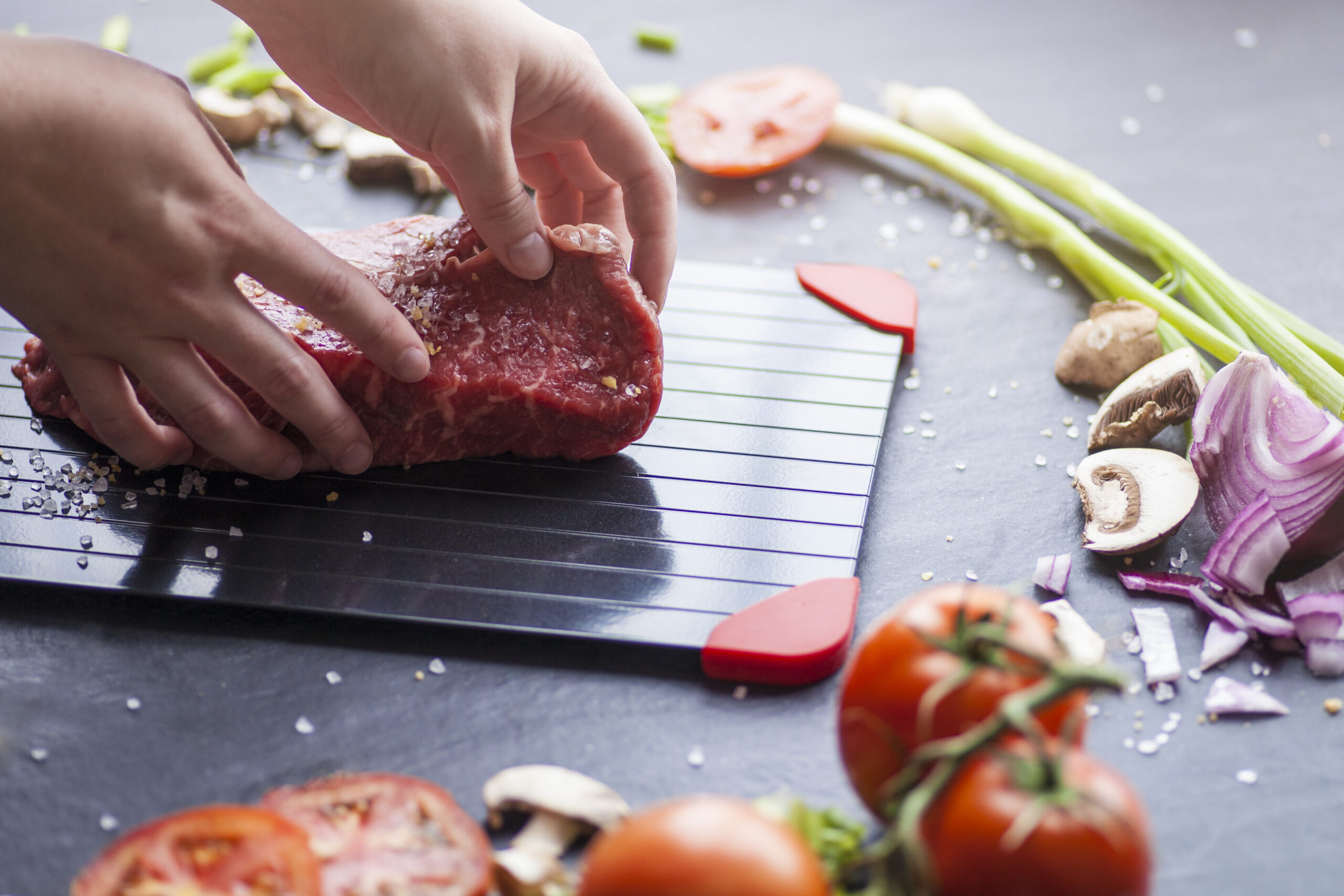 Hands placing raw meat on defrosting tray. The tray is surrounded by chopped food ingredients.