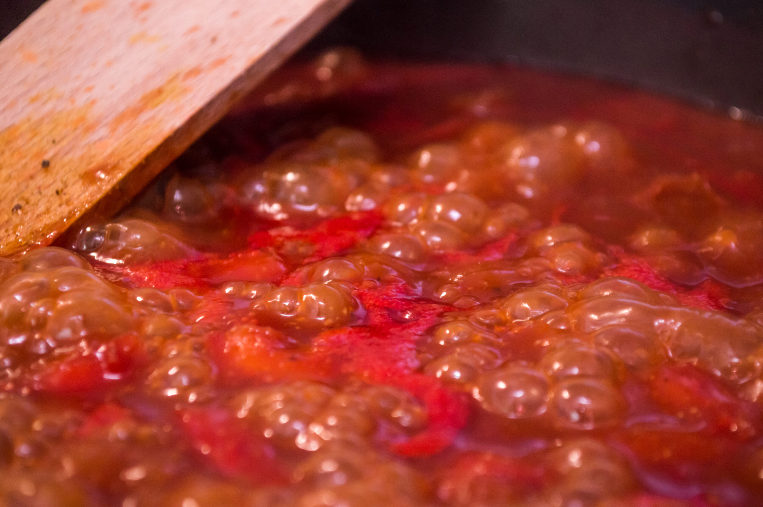 Some bubbling homemade pasta sauce, simmering to reduce and to thicken. A wooden spatula is visible in the background.