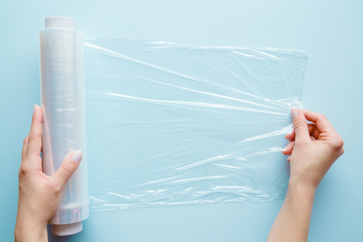 Woman's hand using a roll of transparent polyethylene food film for packing products on the pastel blue table. Empty place for text or logo.