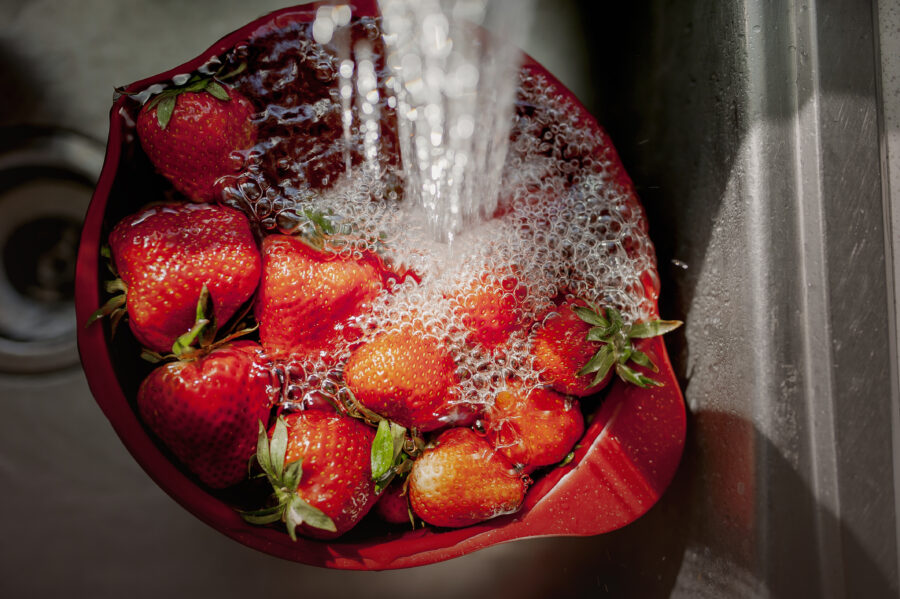 Large group of strawberries floating in a red bowl running under the kitchen faucet