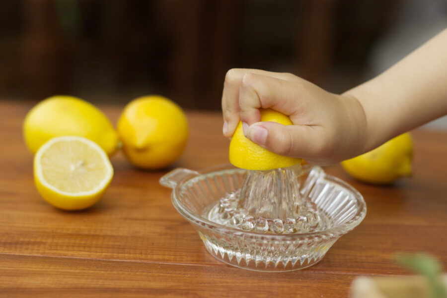 Little hand squeeze lemon juice with a lemon juicer glass on the wood table in the kitchen.