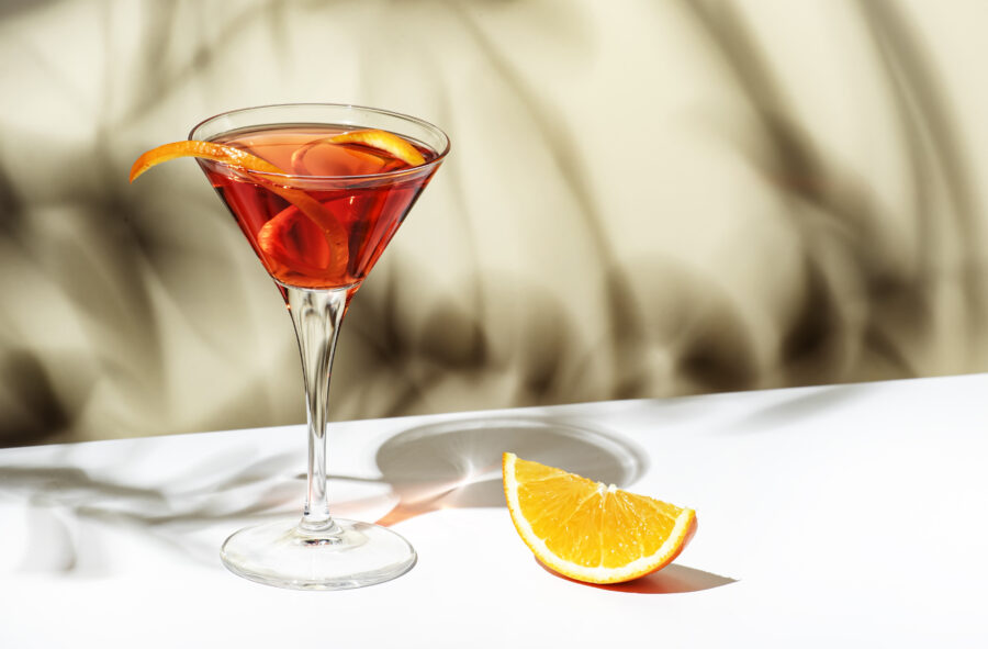 Martinez alcoholic cocktail drink with red vermouth, liqueur, orange bitter, zest and ice. Light beige background, hard light, shadow pattern