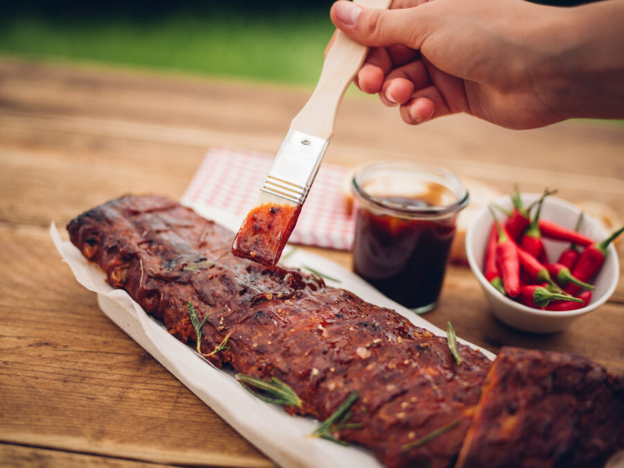 Person's hand holding a brush and painting barbecue sauce onto a rosemary garnished rack of ribs on a wooden table with a bowl of red chillies nearby