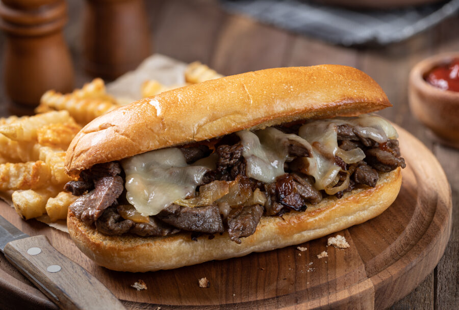Philly cheesesteak sandwich made with steak, cheese and onions on a toasted hoagie roll with french fries on a wooden board