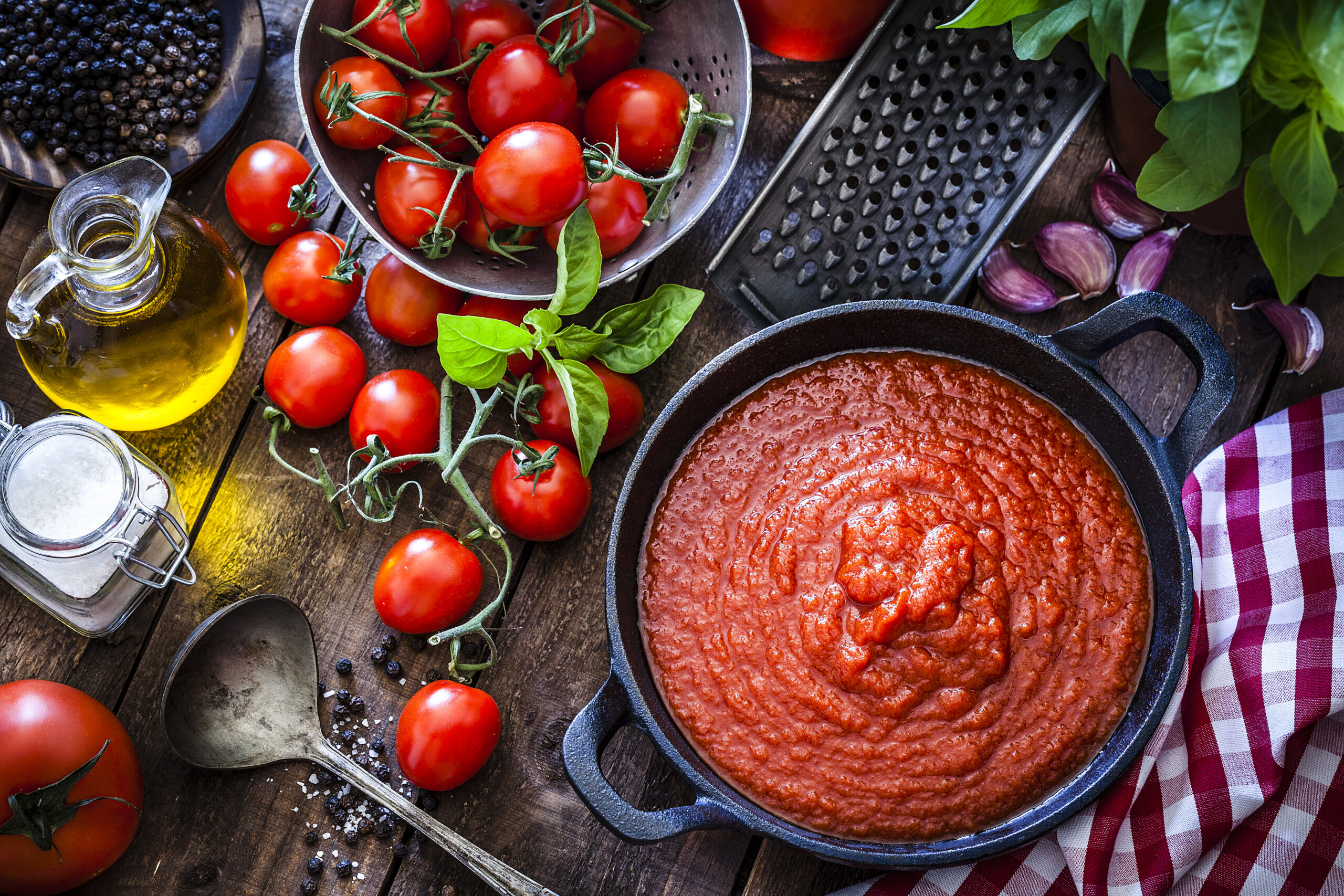 Preparing tomato sauce at home. Top view of a rustic wooden table filled with fresh ingredients for preparing tomato sauce at home. Ingredients for preparing tomato sauce are fresh ripe tomatoes, basil, olive oil, garlic, salt and pepper. The ingredients are scattered on the table all around a cast iron pan filled with prepared tomato sauce. Predominant color is red. DSRL studio photo taken with Canon EOS 5D Mk II and Canon EF 100mm f/2.8L Macro IS USM