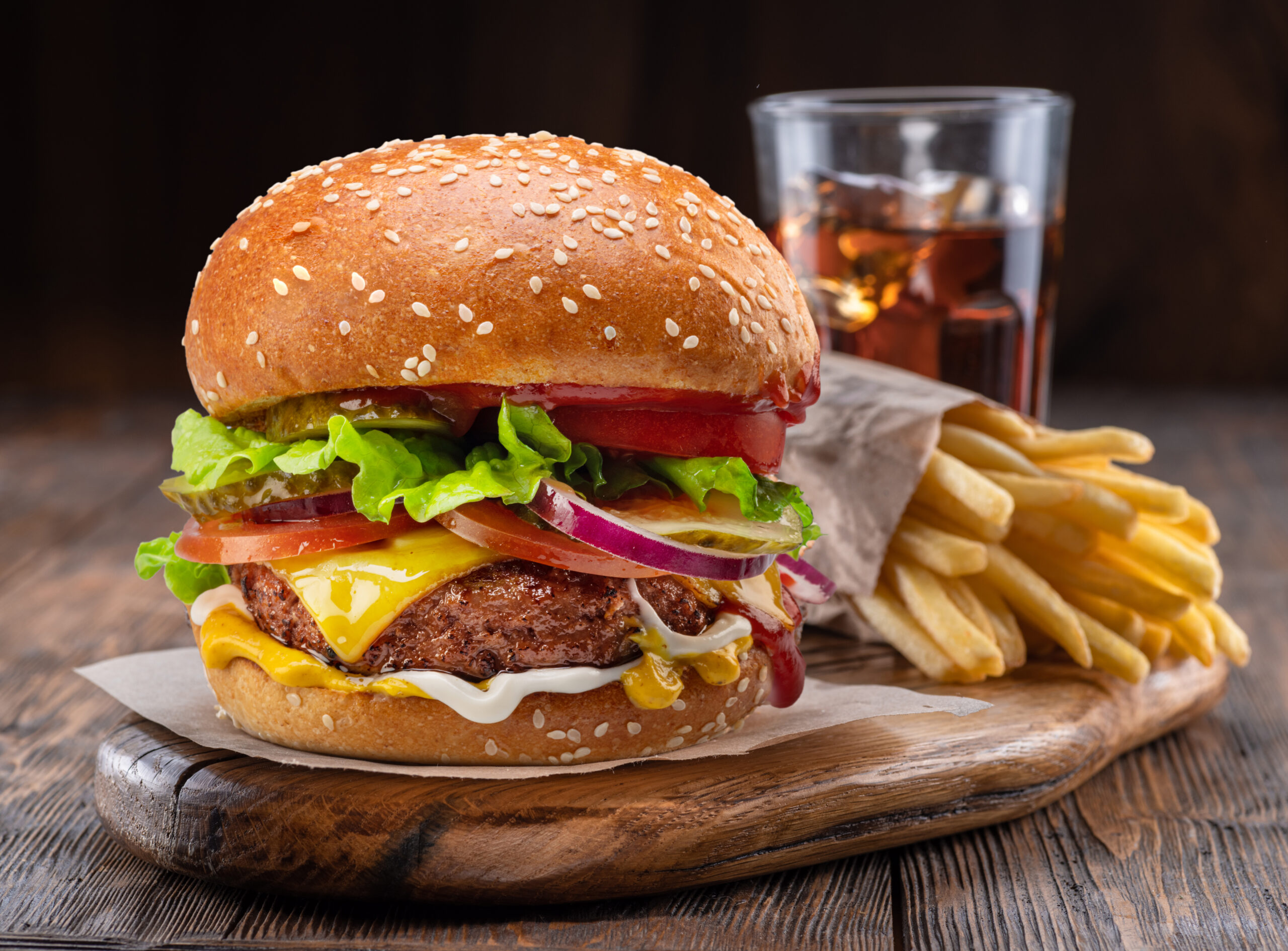 Tasty cheeseburger or hamburger, glass of cola and french fries on wooden tray close-up.